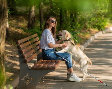 Kör genç bir kadın bankta otururken rehber köpeğe sarılıyor.