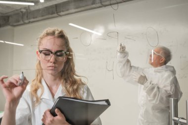 Kadın bir kimyager cama bir formül yazar. Koruyucu giysili yaşlı bir beyaz adam testler yapıyor.