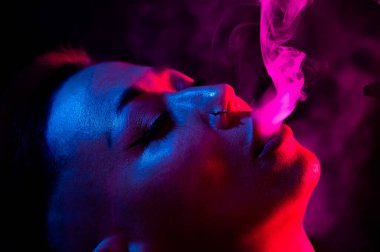 Tıraşlı şakakları olan Asyalı bir kadın neon ışıkta sigara içiyor.