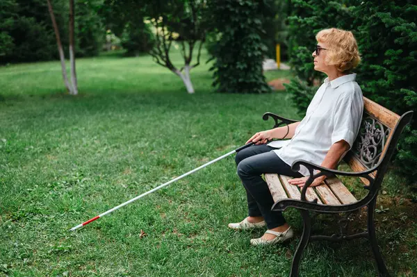 An elderly blind woman sits on a bench in the park
