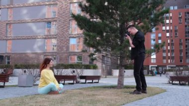 Beyaz kadın çimlerin üzerinde otururken poz veriyor. Bir adam bir mankenin fotoğrafını çeker ve takla atar.