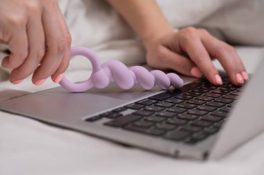 Yatakta yatarken dizüstü bilgisayarın yanında leylak anal boncuklar tutan kadın.