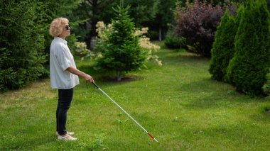 Yaşlı, kör bir kadın, elinde değnekle parkta yürüyor.