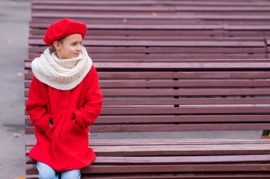 Kırmızı paltolu ve bereli gülümseyen beyaz kız bankta tek başına oturuyor.