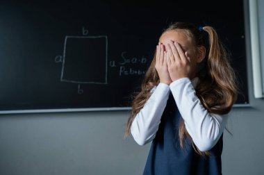Kafkasyalı kız tahtada duruyor ve elleriyle yüzünü kapayarak ağlıyor. Liseli kız ders için hazırlanmadı.