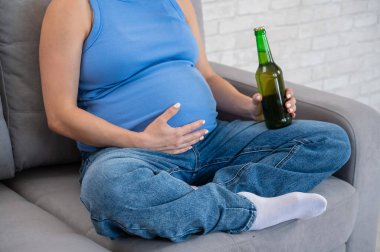 Suratsız hamile kadın kanepede otururken bira içiyor.