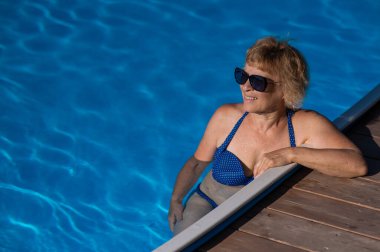 Güneş gözlüklü yaşlı bir kadın havuzda yüzüyor. Emeklilikte tatil