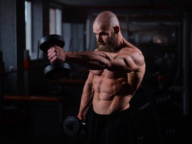 Spor salonunda dambıllarla egzersiz yapan kaslı üstsüz bir adam.