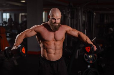 Kaslı üstsüz bir adam spor salonundaki dambıllarla dambılları kaldırır.
