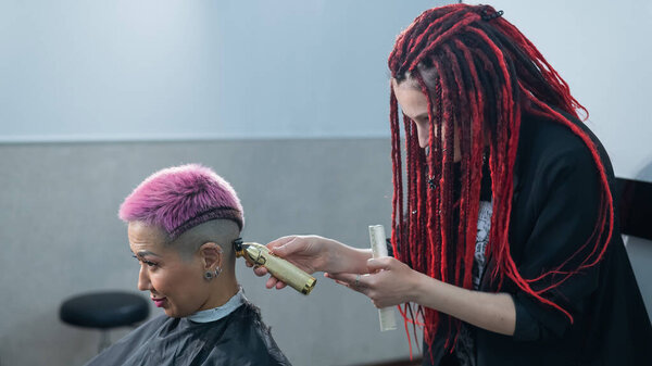 Парикмахер бреет храм клиентки. Азиатка с короткими розовыми волосами в парикмахерской