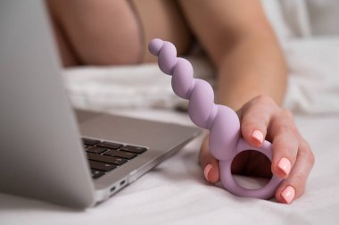 Yatakta yatarken dizüstü bilgisayarın yanında leylak anal boncuklar tutan kadın.