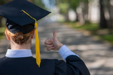 Mezuniyet elbisesi giymiş beyaz bir kadının arka planda baş parmağı açık havada görünüyor.