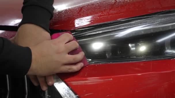 主人把乙烯薄膜涂在一辆红色汽车的前灯上 工人用刮板保护膜更精细地抚平手的布景 — 图库视频影像