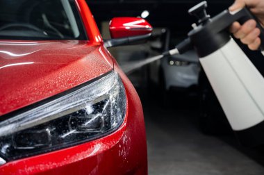 Bir adam kırmızı bir arabanın farlarını spreyle yıkıyor.