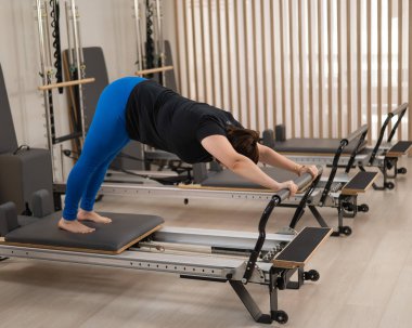 Aşırı kilolu beyaz kadın bir reformcu üzerinde pilates egzersizi yapıyor.