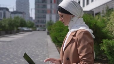 Başörtüsü takmış genç bir kadın bir bankta oturur, dizüstü bilgisayarda çalışır ve arkadaşına el sallar.