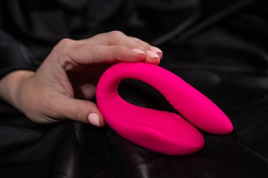 Bir kadın siyah ipek çarşafın üzerinde yatarken pembe kıvrımlı vibratör tutuyor.