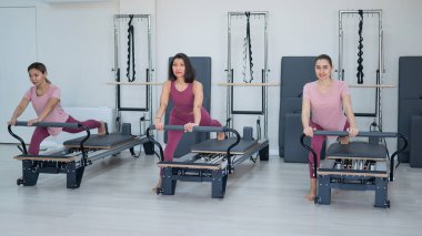 Three Asian women in pink sportswear do exercises on the reformer machine. Pilates classes