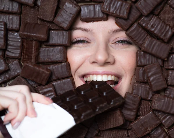 The face of a caucasian woman surrounded by chocolates. The girl eats a bar of chocolate
