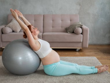 Hamile kızıl saçlı kadın evde fitness topu egzersizleri yapıyor.