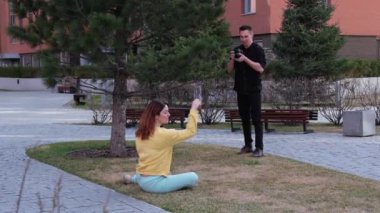 Beyaz kadın çimlerin üzerinde otururken poz veriyor. Bir adam bir mankenin fotoğrafını çeker ve takla atar.