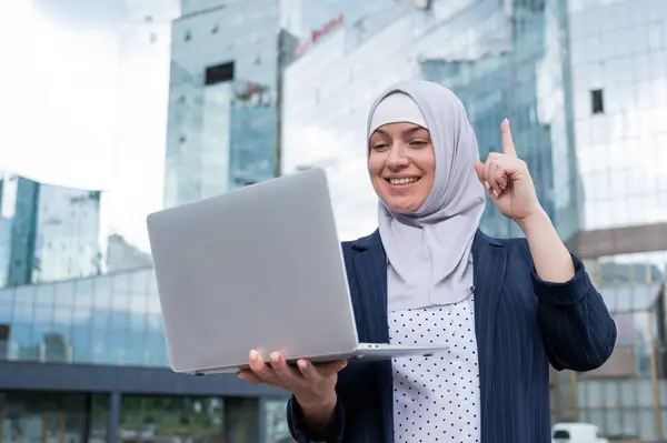 Business woman in hijab and suit is using a laptop and holding her index finger up outdoors