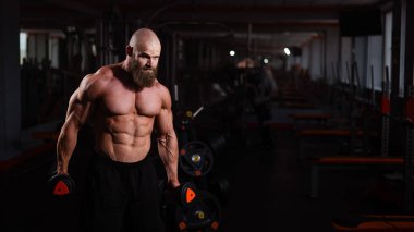 Spor salonunda dambıllarla egzersiz yapan kaslı üstsüz bir adam.