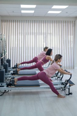 Three Asian women in pink sportswear do exercises on the reformer machine. Pilates classes