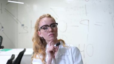Tıbbi elbise giymiş beyaz bir kadın şeffaf bir duvarda formülleri düşünüp nihayete erdiriyor.