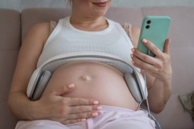 Hamile bir kadın, çocuğu için akıllı telefondan müzik açar. Kulaklıklar karnında.