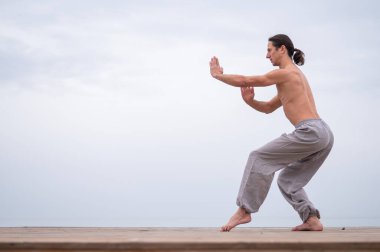 Beyaz bir adam, çıplak gövdesiyle deniz kıyısında wushu yapıyor.