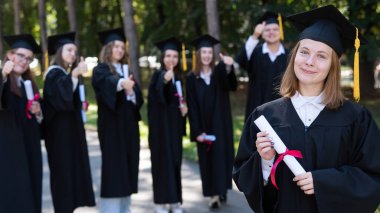 Mezuniyet elbisesi giymiş bir grup mutlu öğrenci dışarıda. Ön planda elinde diploması olan genç bir kız.