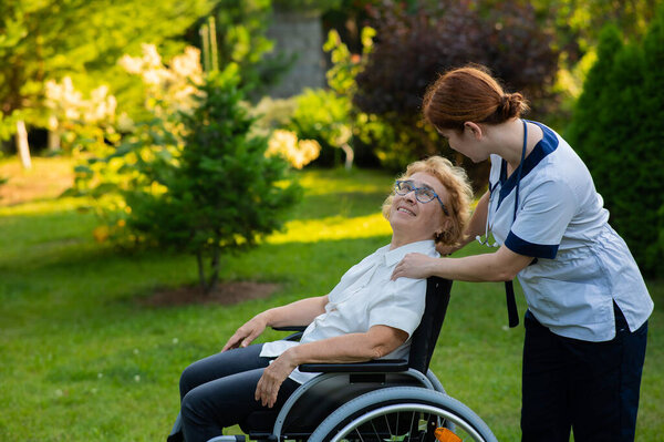 A nurse holds an elderly caucasian woman in a wheelchair by the hand as support. Nurse walks with a patient in the park