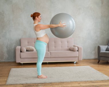 Hamile kızıl saçlı kadın evde fitball ile kol egzersizi yapıyor.