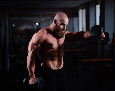 Spor salonunda dambıllarla egzersiz yapan kaslı üstsüz bir adam.