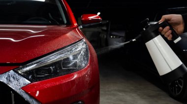 Bir adam kırmızı bir arabanın farlarını spreyle yıkıyor.