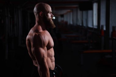 Kaslı kel bir adam karanlıkta üstsüz poz verir. Vücut geliştiren biri spor salonunda vücudunu gösterir.