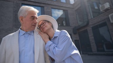 Şapkalı ve gözlüklü bir kadın beyaz ceketli yaşlı bir adama arkadan sarılıyor. Olgun insanların romantik ilişkileri