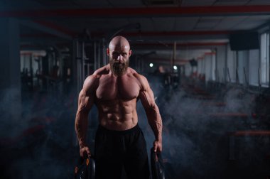 Ağırlık plakası tutan kaslı, kel, üstsüz bir adam. Vücut geliştiren biri spor salonunda vücudunu gösterir.