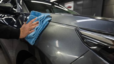 Bir adam gri bir arabanın yüzeyini mikrofiber bir bezle siliyor.