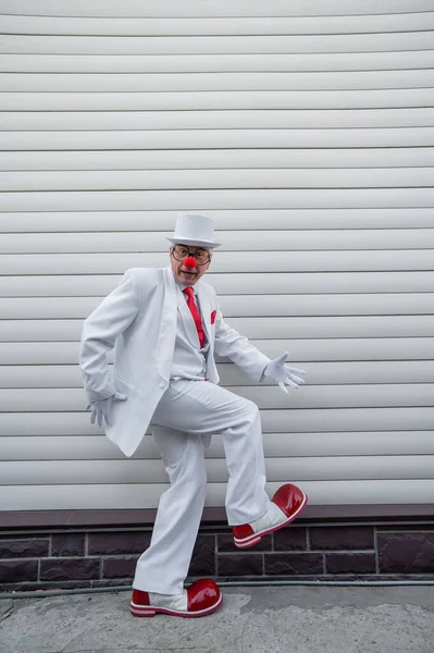 An elderly man in a white suit, huge boots and a clown nose walks funny