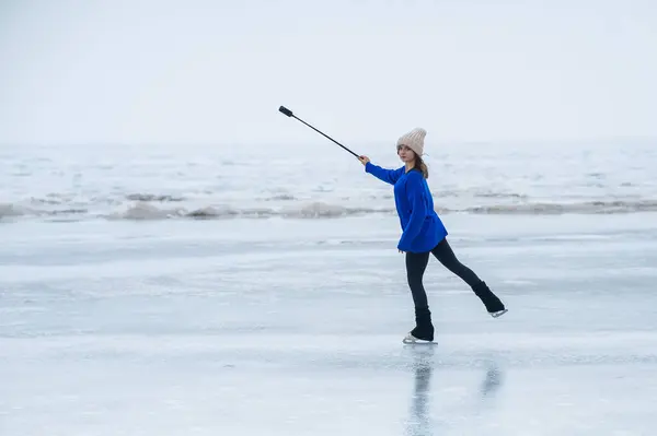 A caucasian woman is skating on a frozen lake holding a selfie stick in her hands. The figure skater films her skating