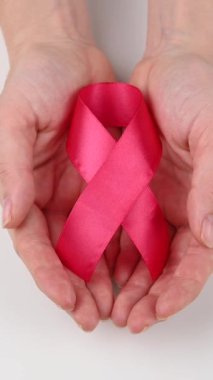 Beyaz arka planda pembe kurdeleli kadın ellerinin yakın çekimi. Göğüs kanseriyle mücadelenin sembolü. Dikey video