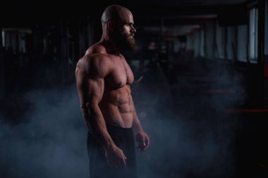 Kaslı kel bir adam karanlıkta dumanın ortasında üstsüz poz verir. Vücut geliştiren biri spor salonunda vücudunu gösterir.