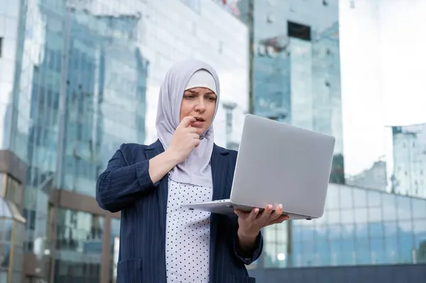 Thoughtful business woman in hijab and suit is holding a laptop outdoors