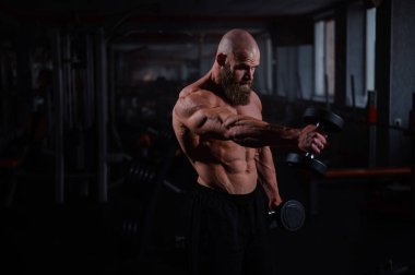 Spor salonunda dambıllarla egzersiz yapan kaslı üstsüz bir adam.