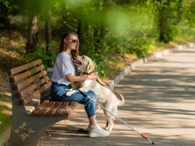 Kör genç bir kadın bankta otururken rehber köpeğe sarılıyor.