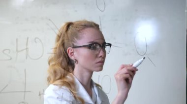 Tıbbi elbise giymiş beyaz bir kadın şeffaf bir duvara formüller yazıyor.