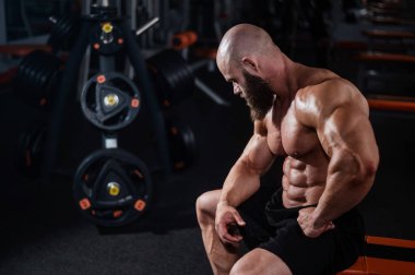 Kısa şortlu, kaslı, kel bir adam antrenmandan sonra bankta dinleniyor. Vücut geliştirici spor salonunda vücudunu gösteriyor.