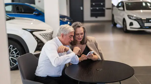 Dojrzała Para Siedzi Salonie Samochodowym Patrzy Smartfona — Zdjęcie stockowe